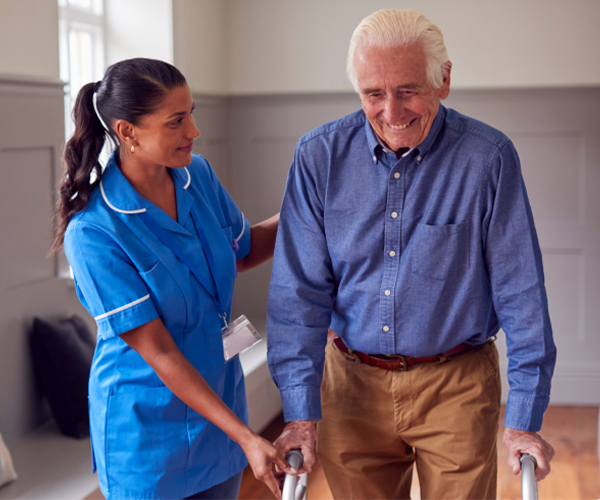 A caregiver in blue scrubs happily helping an elderly man walk.